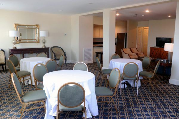 A hotel room suite has three small round tables set with tablecloths and chairs around them. A small kitchen and living room space are to the back side of the space.