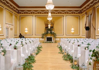 Our gold ballroom is set with white chairs with greenery accents in rows for a wedding ceremony. A large ornate mirror is int he center of he room and a piano sits off to the left hand side.