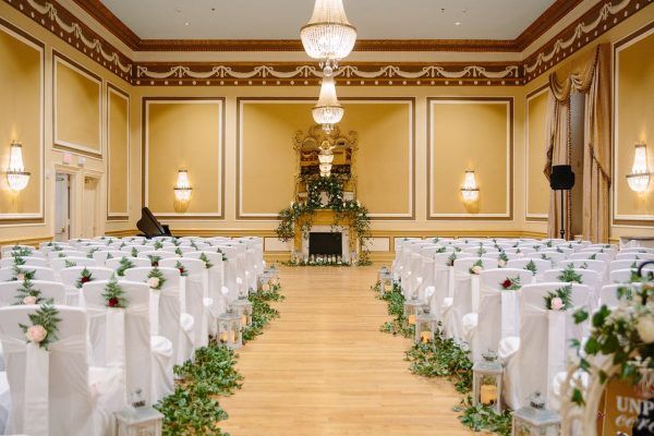 Our gold ballroom is set with white chairs with greenery accents in rows for a wedding ceremony. A large ornate mirror is in the center of he room and a piano sits off to the left hand side.