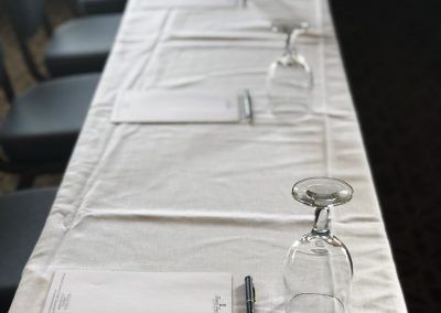 A long rectangular table has a pad of paper, pens and empty water glasses set.