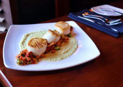 A swuare white plate has three lightly browned scallops that sit on top of a vegetable relish and light green hummus spread.