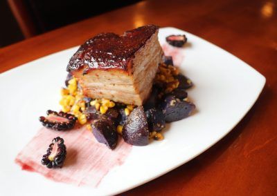 A thick cut of pork belly that shimmers with glaze sits on top of a yellow corn, purple potato and blackberry medley.