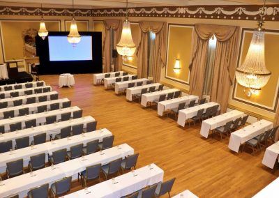 An aerial view of the gold ballroom is set with multiple long rectangle tables with blew chairs. They are facing a large projector screen and a small stage with a podium is to the side.