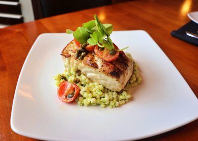 A thick cut of a white fish is lightly seared to a brown hue and small round grains of couscous sit beneath the fish. Red cherry tomatoes and green leaves garnish the top of the fish,