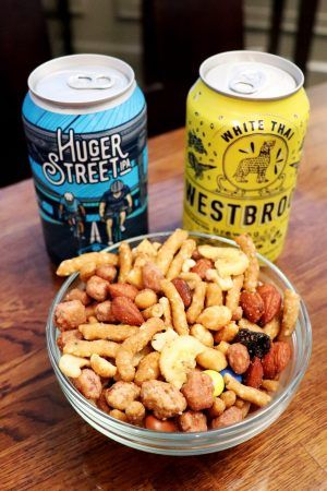 A small bowl of trailmix and two cans of beer sit on a table.