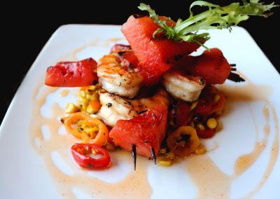 A white square plate with shrimp and watermelon on skewers sit on top of peppers and corn.
