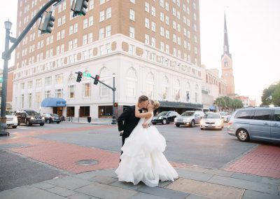 A bride and groom embrace in a kiss on the corner on the street with the hotel in background as traffic goes by