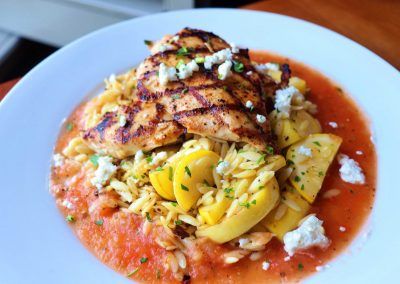 Grilled chicken on a bed of orzo rice with yellow squash and a red sauce in placed on top of a round white plate.