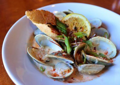 A small white bowl is filled with littleneck clams that are opened and a garnished with green onion and a lemon. Two slices of bread are set in the middle of the clams.