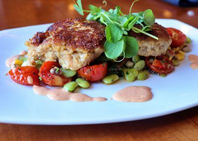 Two seared crabcakes sit on top of a bed of cherry tomatoes, green edmame beans and yellow corn. A light orange sauce isaround the dish and green leaves garnish the tops of the crabcakes.
