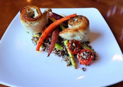 Two roll ups of a white fish filet are placed on top of small quinoa grains with purple and orange carrots and red tomatoes.