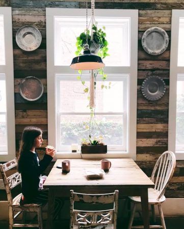 A small girl sits at a table next to a large window and is eating a pastry.