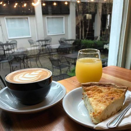 A cup of coffee, glass of orange juice and a piece of quiche sit at the edge of a table and looks out into a window.