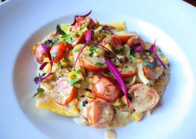 Ravioli with a cream suace with bright red cherry tomatoes and yellow corn is garnished with bright purple and green leaves.