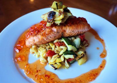 A round white plates has a bed of rice with green and yellow squash with red peppers. A seared piece of pink salmon sits on top of the medley of vegetables and a red pepper jelly is circled around the edge of the dish.