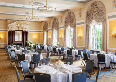 Our colonial ballrooms offers large windows letting in tons of natural light. The rooms is set with multiple large round tables with blue chairs seated around each table. Small floral centerpieces sit in the middle of the tables and an ornate fireplace is in the center of the back wall. Large antique chandeliers hang from the ceiling.