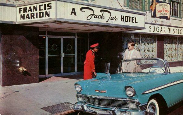 An old postcard that has an image of a bellman in a red suite opening the door to a blue car for a man and women to get out. The hotel's front doors are in the background.