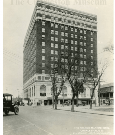A black and white image of the hotel from the street. A few cars are parked in front of the hotel and to the side. A couple of people are standing or walking at the corner of the hotel. A few bare trees are lining the park that sits in front of the building.
