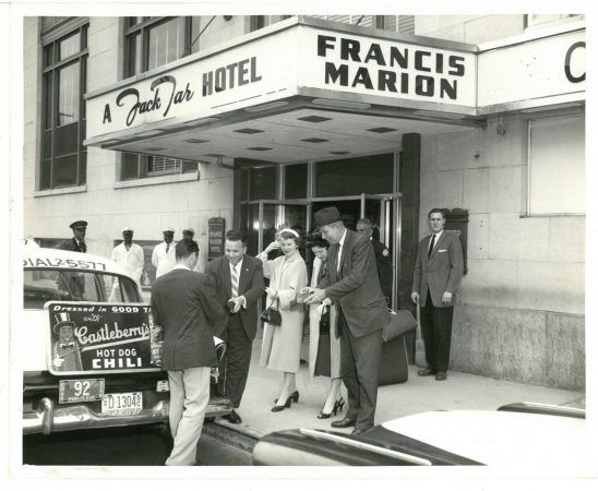 A black and white image o a group of people getting our of a taxi in front of the hotel and paying their cab driver.