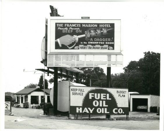 A black and white images of a billboard from a street view. The billboard reads 'The Francis Marion Hotel Proudly Presents Beef on a dagger! in the Swamp Fox Room. New! The trophy room'. A hand holding a dagger with four pieces of meat takes up the center of the billboard.
