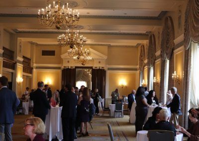 A gathering in a ballroom space. People are mingling and walking around the ballroom that is set with different sized tables.