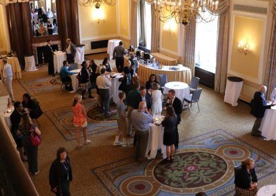 A gathering in a ballroom space. People are mingling and walking around the ballroom that is set with different sized tables.