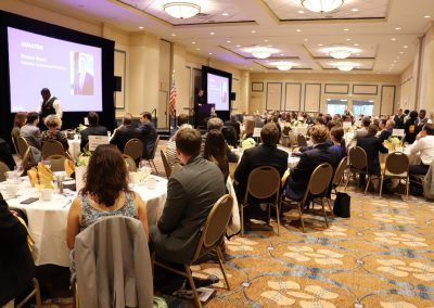 A ballroom in the hotel has a conference taking place and multiple round tables are filled with people listening o a presentation by a man.