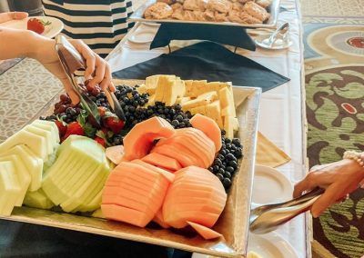 A buffet with a large plate of fruit and two people are using tongs to plate their fruit. A lady behind is grabbing a pastry off another plate and a plate of bagels sits at the end of the table.