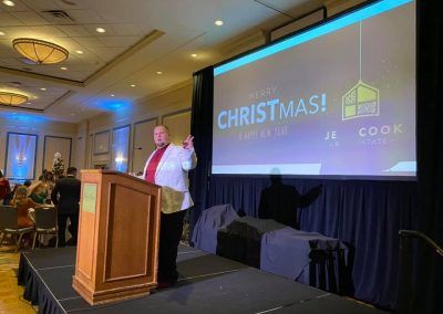 A man stands at a wooden podium on a stage is giving a speech and points off into the distance. A screen is behind him that reads merry Christmas and happy new year