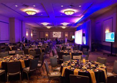 A ballroom space has purple and blue lighting and is set with multiple round tables with salas, dessert and drinks already on the tables. A projector places the words Health Care Heroes on the back wall.