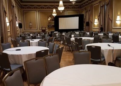 A ballroom space is set with multiple round tables that are bare. A large blank screen is in the back.