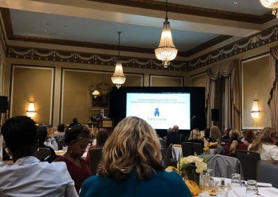 A conference is taking place in a ballroom. People are sitting at tables looking up at the speaker at the front of the room and a screen is projecting.