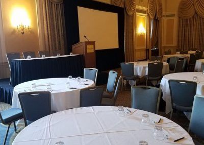 A ballroom space that is set for a conference with round tables and a stage that has a podium, a large screen and a rectangle table.