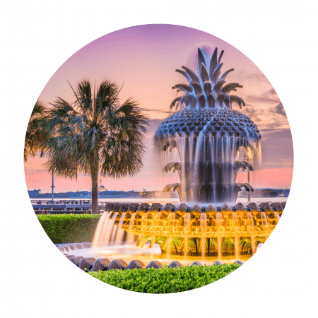 A fountain shaped like a pineapple has water cascading down it. A palm tree is in the background and the sun is setting.