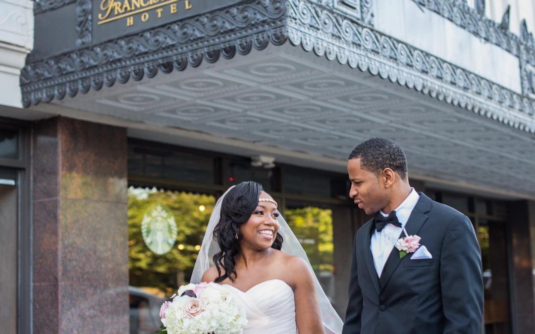 A bride and groom looking at each other and smiling are walking in front of the hotel.