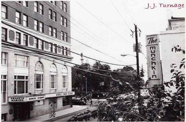 A black and white images of the hotel building outside. A sig to the right reads The Munchkin. There is the park in the background and a few people walk on the streets.