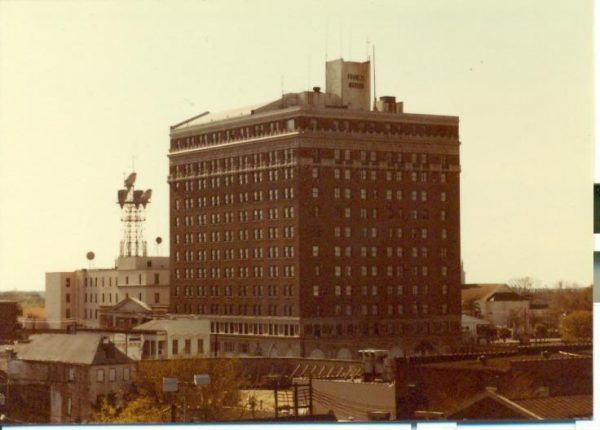 A sepia toned image of the exterior of the hotel from the 1980s. Buildings sit below it as the hotel rises twelve stories tall.