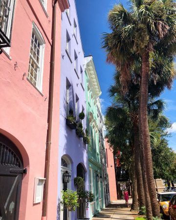 Pink, purple, blue color houses line a street as a lady in a pink outfit walks on the sidewalk towards the camera. Tall palm trees line the other side of the sidewalk and the sky is bright blue and its sunny out.