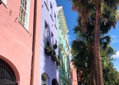 Pink, purple, blue color houses line a street as a lady in a pink outfit walks on the sidewalk towards the camera. Tall palm trees line the other side of the sidewalk and the sky is bright blue and its sunny out.