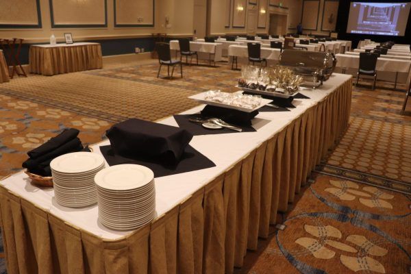 A long table with plates and silverware. Two bigger plates with wrapped food and a silver warming chaser sits in the middle of the table. The table sits in a large ballroom that has blue and gold patterned carpet.