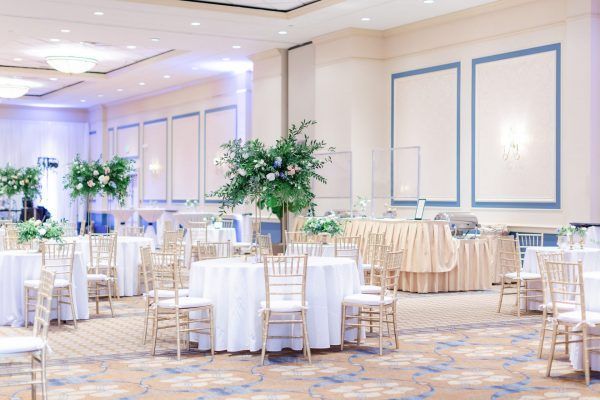 A brightly lit ballroom space with multiple round tables and large green floral displays. A bar with gold draped fabric is against the wall.