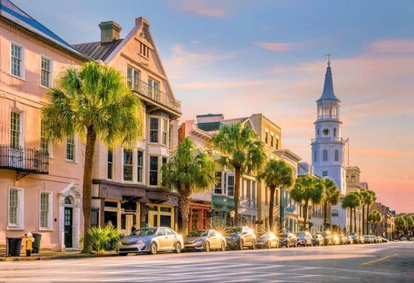 A street in Charleston is lined with cars and historic buildings and palm trees. The sun is setting and shines brightly against the cars and trees.