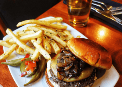 A burger with sautéed mushrooms and a white cheese. Yellow crispy fries sit next to the burger and green okra and pickles garnish the plate.