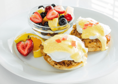 A plate of eggs benedict with yellow hollandaise sauce and a cup of fruit filled with red strawberries, yellow pineapple and blueberries.