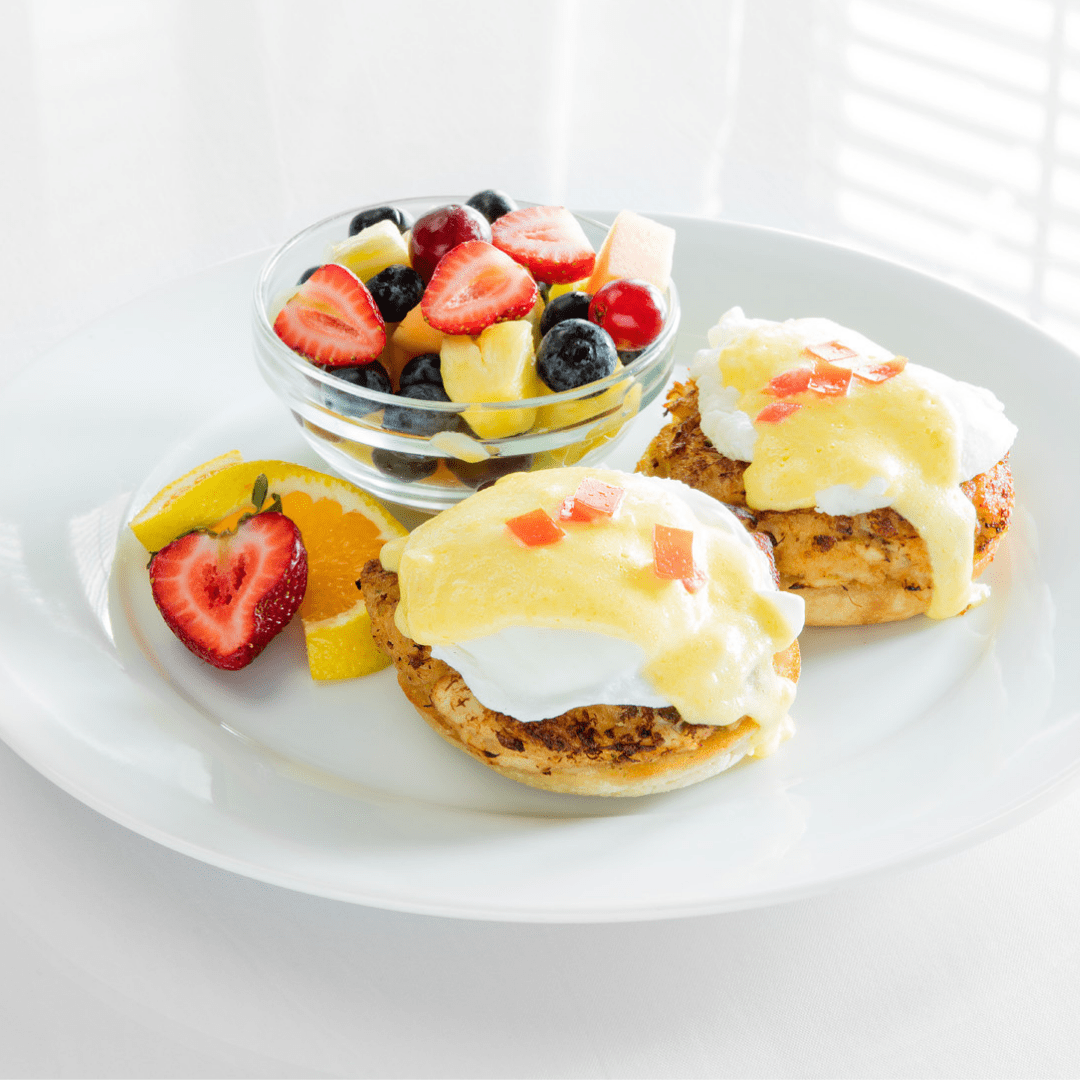 A plate of eggs benedict with yellow hollandaise sauce and a cup of fruit filled with red strawberries, yellow pineapple and blueberries.