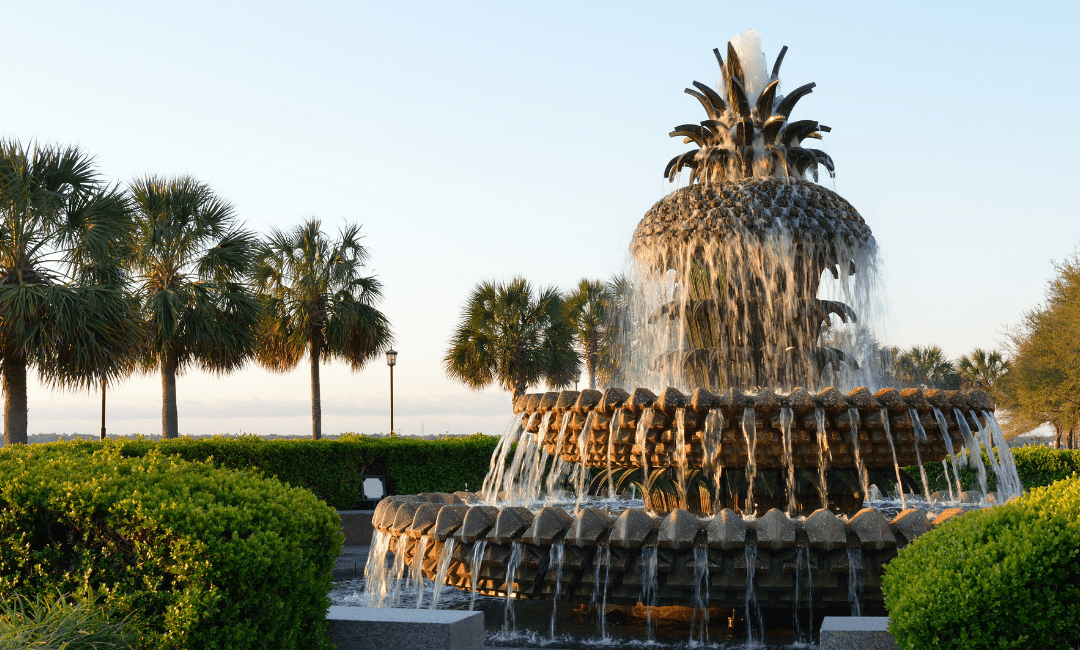 The pineapple fountain has water cascading down. Palm trees line the park the fountain sits in.