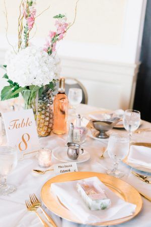 A table is set for a wedding reception. A floral centerpiece sits int eh middle with white and pink florals and a pineapple accent next to it. A gold plate and gold silverware are in the center.