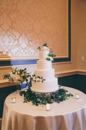 A white wedding cake with green and white floral accents sit on top of a round table with a white tablecloth. A few candles sit around the cake.
