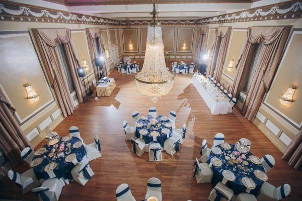 Our Gold ballroom is shown from above. Round tables are scattered throughout with deep blue tablecloths. A long, rectangular table is in the middle for the bridal party to sit at. Gold accents are throughout and a gran chandelier sits in the middle of the room.