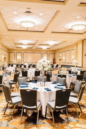 A ballroom is set for a wedding reception. Round ables are set with white tablecloths and blue napkins. Blue chairs sit around the tables. White flowers are in the center of the tables.